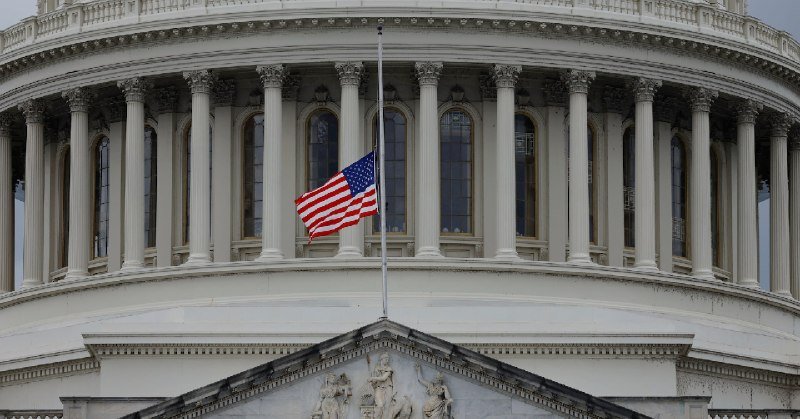 An American flag flies at half-staff on a flagpole in front of a domed government building with classical columns and sculptural reliefs.