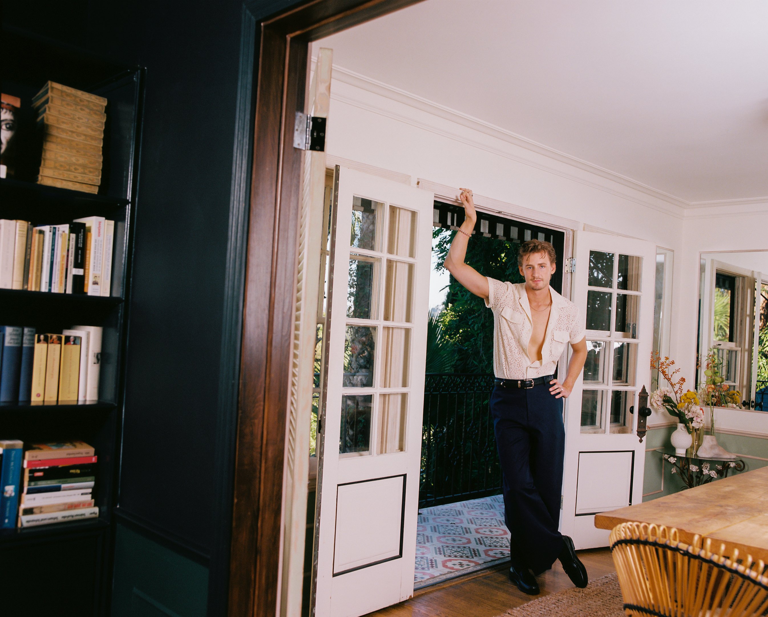 A man in an unbuttoned shirt and dark trousers stands in the doorway of a home, one arm raised above his head, framed by open French doors leading to a garden.