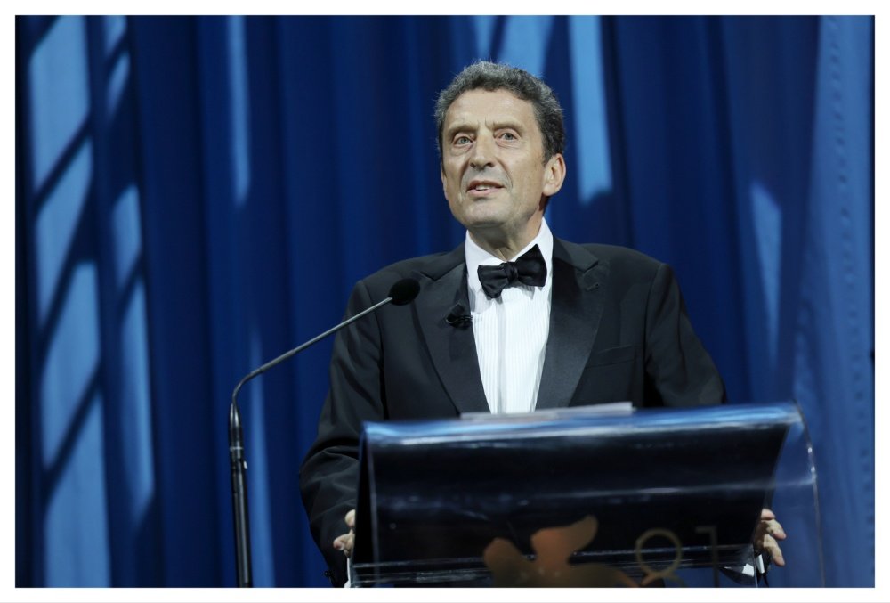 A man in a black tuxedo and bow tie speaks at a clear podium with a microphone, set against a blue curtain backdrop.