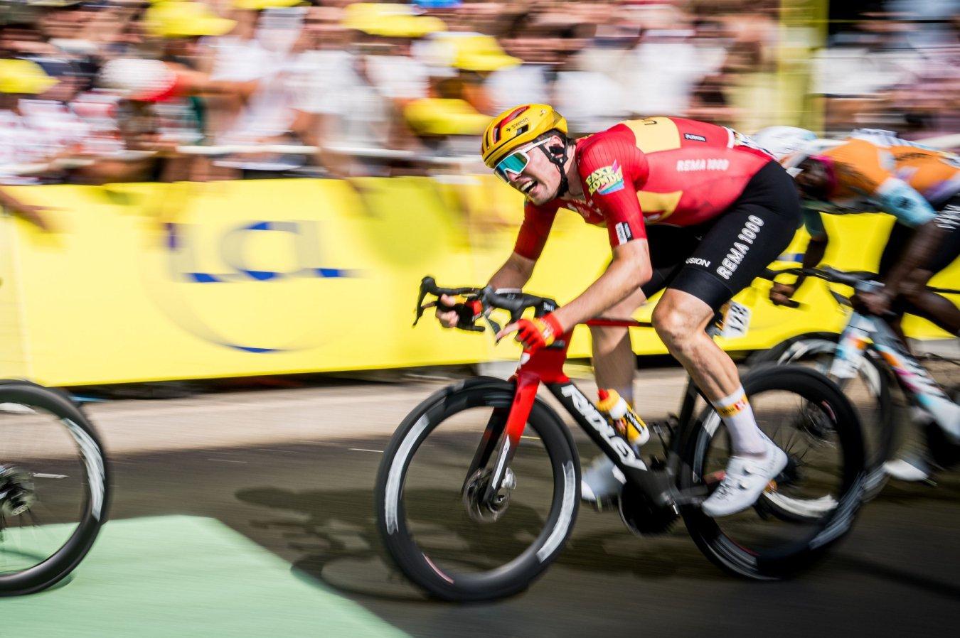 A cyclist in a red jersey and yellow helmet sprints on a Ridley bike past cheering crowds behind yellow barriers.
