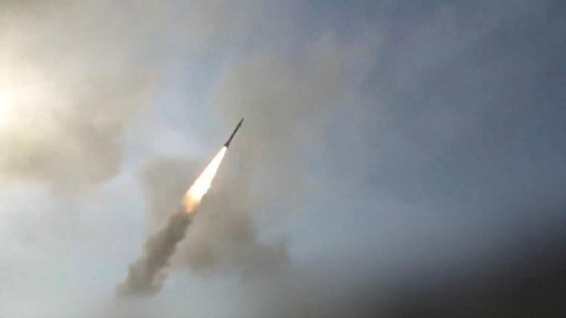 A missile ascends through a cloudy sky, leaving a bright flame and thick smoke trail behind it.