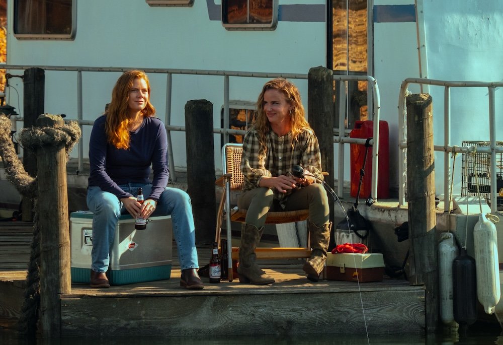 Two women sit on a wooden dock beside a boat, one holding a beer bottle while seated on a cooler and the other holding a fishing rod in a chair.
