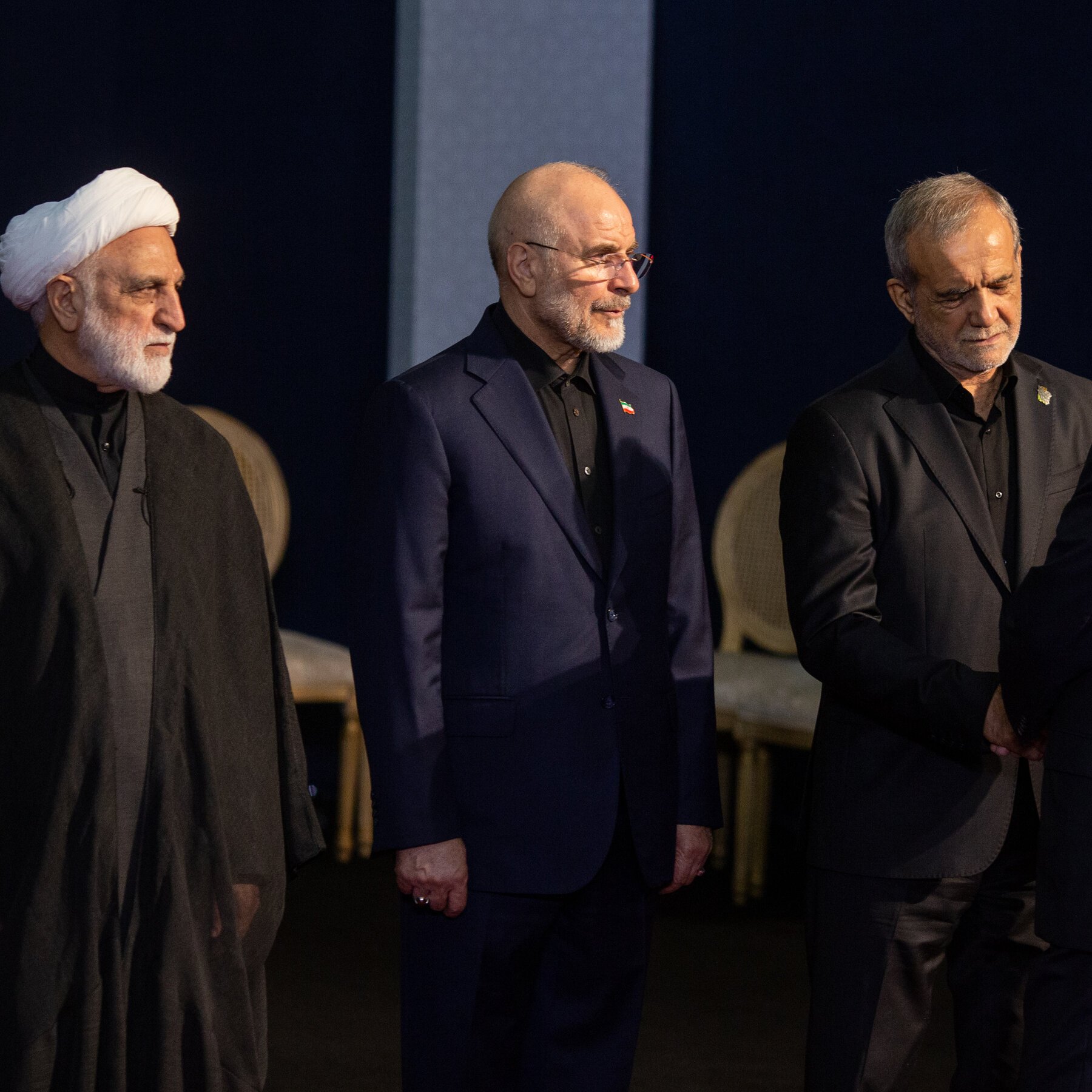 Three men in dark formal attire, including one wearing a white turban and another wearing an Iranian flag pin, stand solemnly against a dark backdrop.