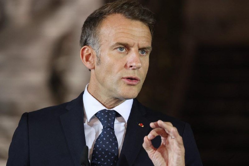 A man in a dark suit, white shirt, and patterned tie gestures with his hand while speaking, with a small pin on his lapel, against a blurred background.