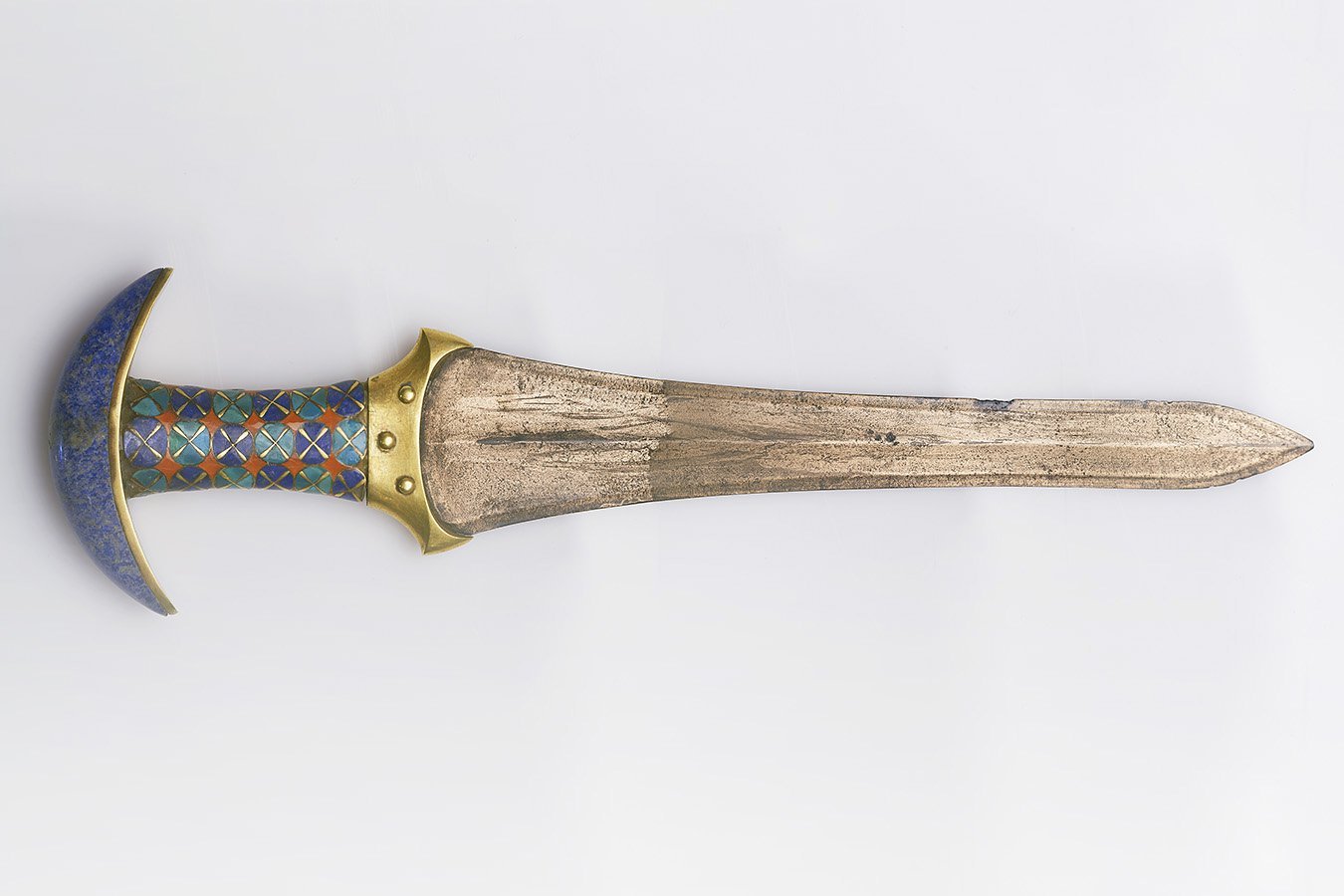 An ornate dagger with a blue crescent-shaped pommel, cloisonné-encrusted handle, and a weathered bronze-colored blade rests against a white background.