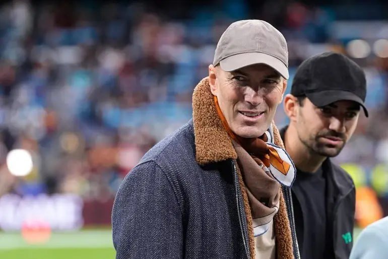 Two men wearing caps and jackets stand smiling in a stadium with a blurred crowd visible in the background.
