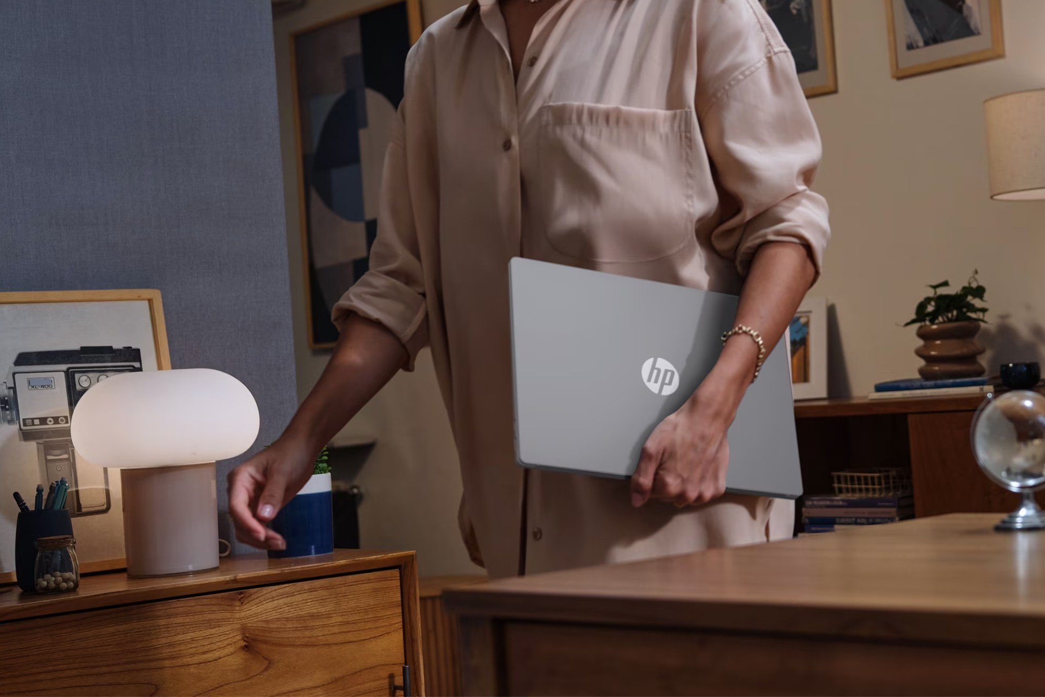 A person in a beige shirt carries an HP laptop away from a wooden desk in a warmly lit, decorated room.