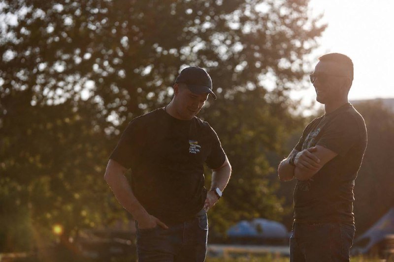 Two men wearing dark t-shirts, baseball caps, and sunglasses stand outdoors at sunset, engaged in conversation.