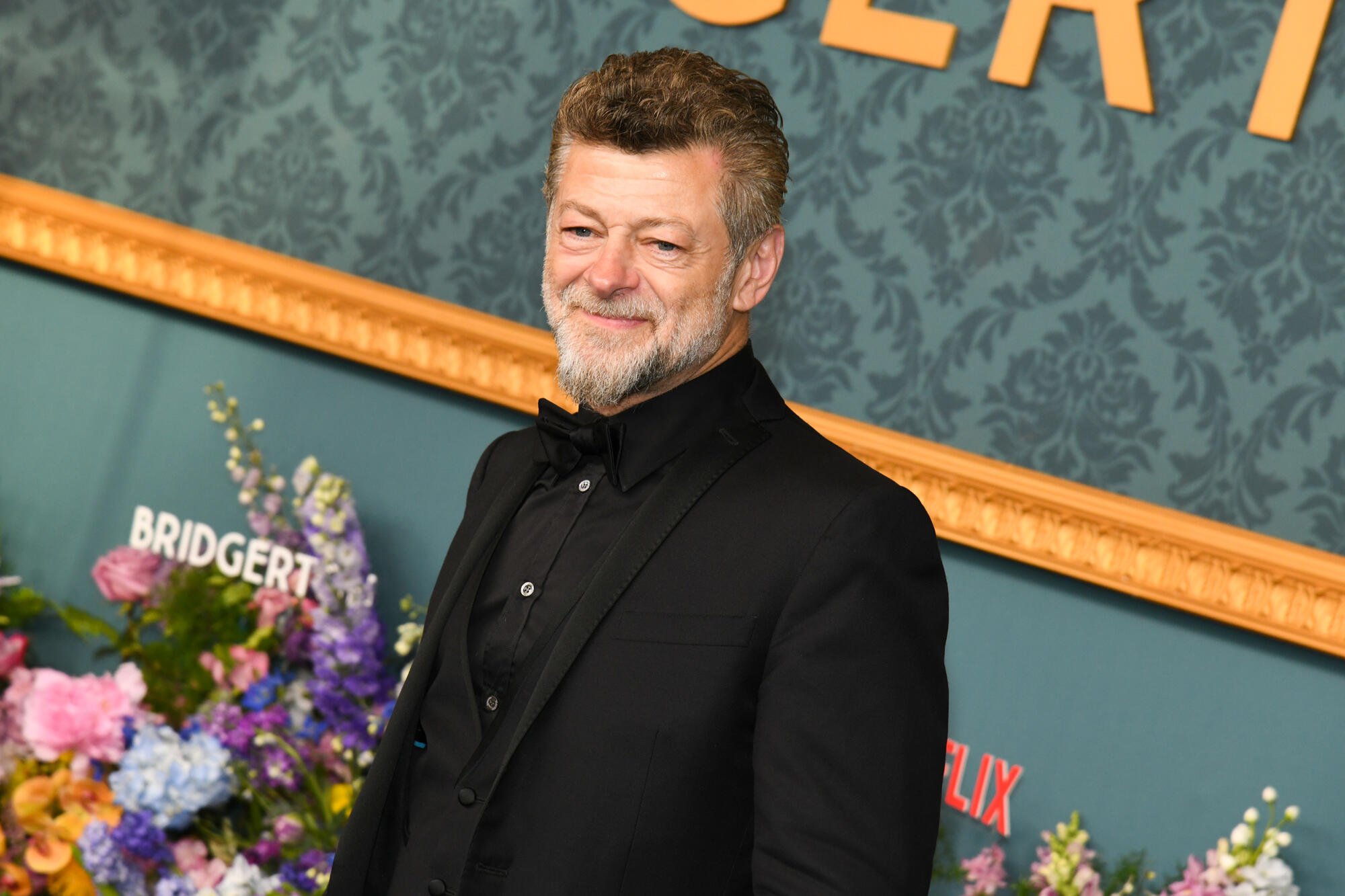 A bearded man in a black tuxedo smiles on a step-and-repeat backdrop reading "Bridgerton" and "Netflix," flanked by floral arrangements.