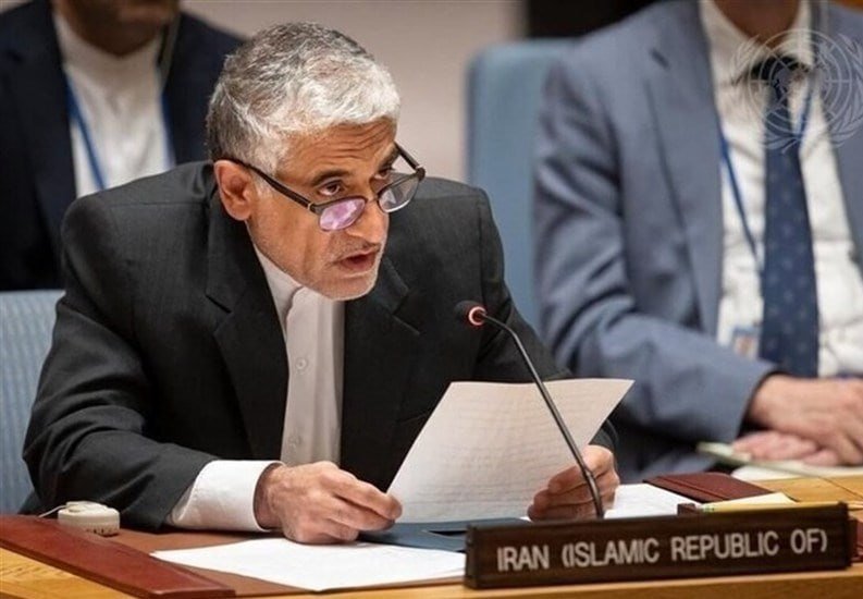 A gray-haired man in glasses and a dark suit reads from a paper while speaking into a microphone at a desk labeled "Iran (Islamic Republic of)."