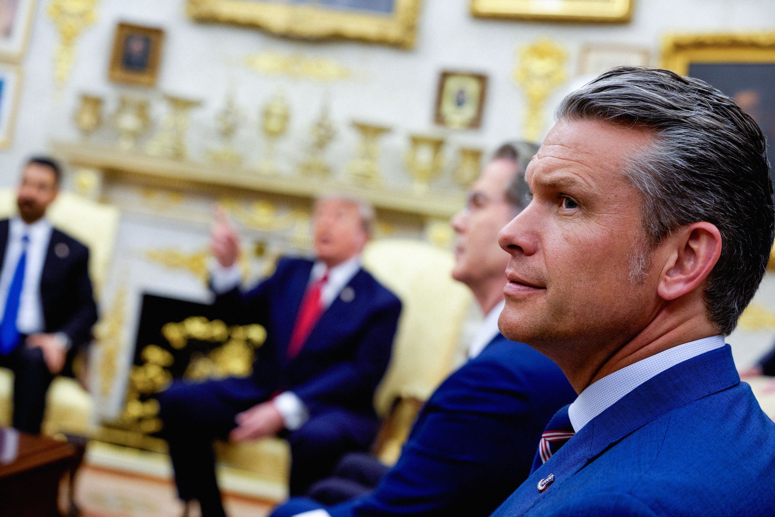 Men in blue suits and ties sit in an ornate room featuring gold-framed artwork and decorative wall panels.
