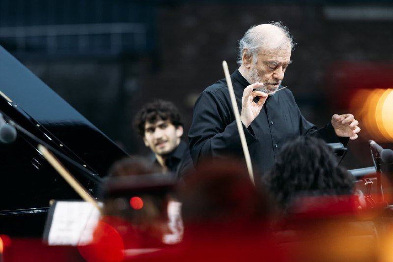 An older bearded conductor in black gestures with a baton, directing musicians seated in an orchestra pit with a grand piano.