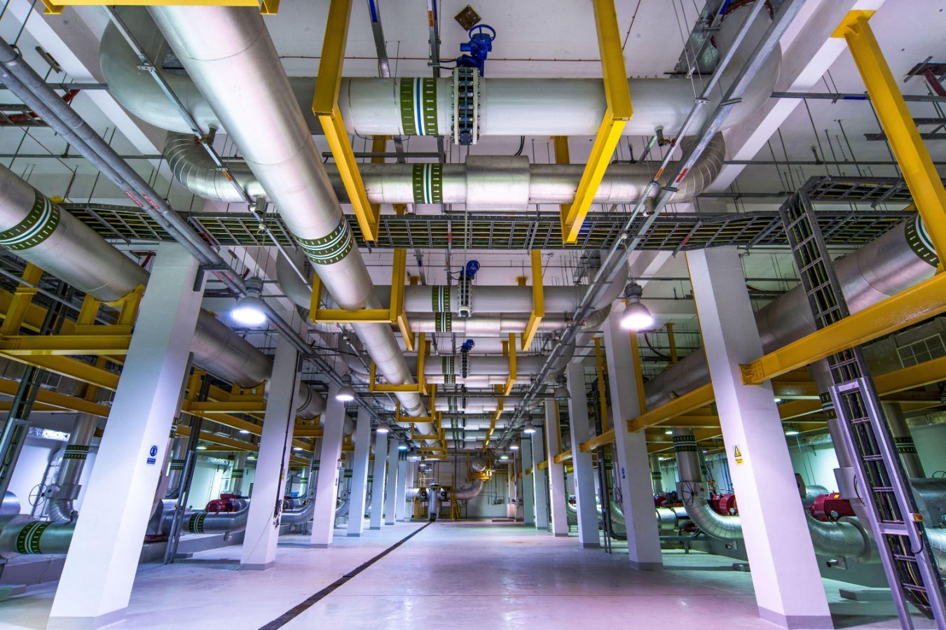 A long industrial facility interior featuring large overhead pipes, yellow support beams, white columns, and metal walkways under bright ceiling lights.