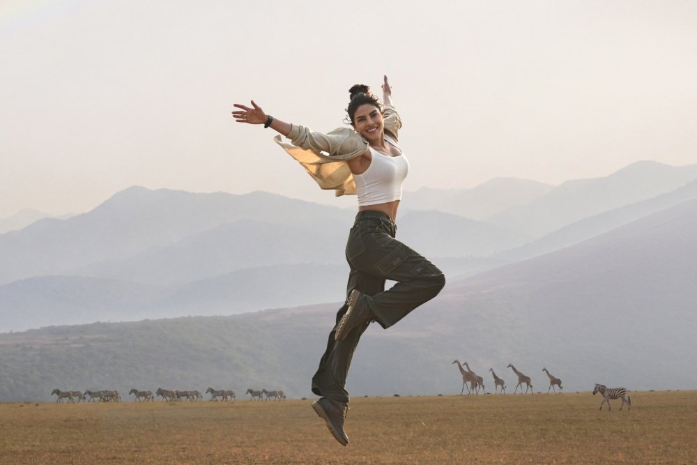 A woman jumps joyfully with arms outstretched against a savanna backdrop featuring a zebra and giraffes walking across the plains below hazy mountain ranges.