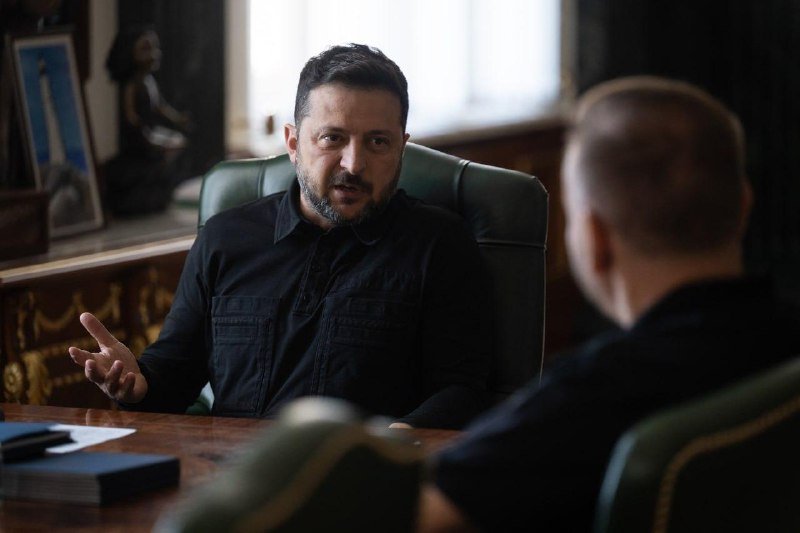 A bearded man in a dark shirt sits at a desk, gesturing with his hand while speaking to another person seated across from him in a formal office setting.