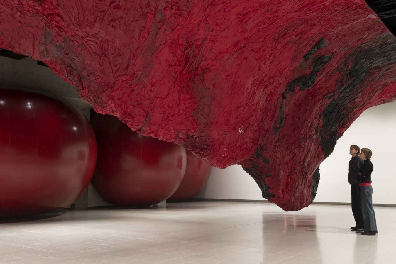 Two people stand in a gallery looking up at a massive red and black sculptural installation suspended above large glossy red spheres.