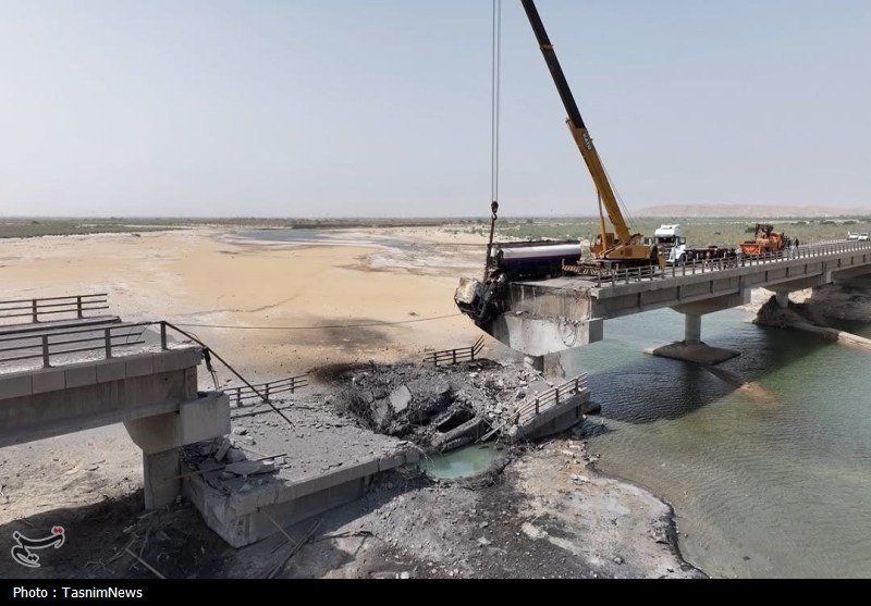 A yellow crane operates on a partially collapsed bridge spanning a river, with workers present and surrounding infrastructure visibly damaged.