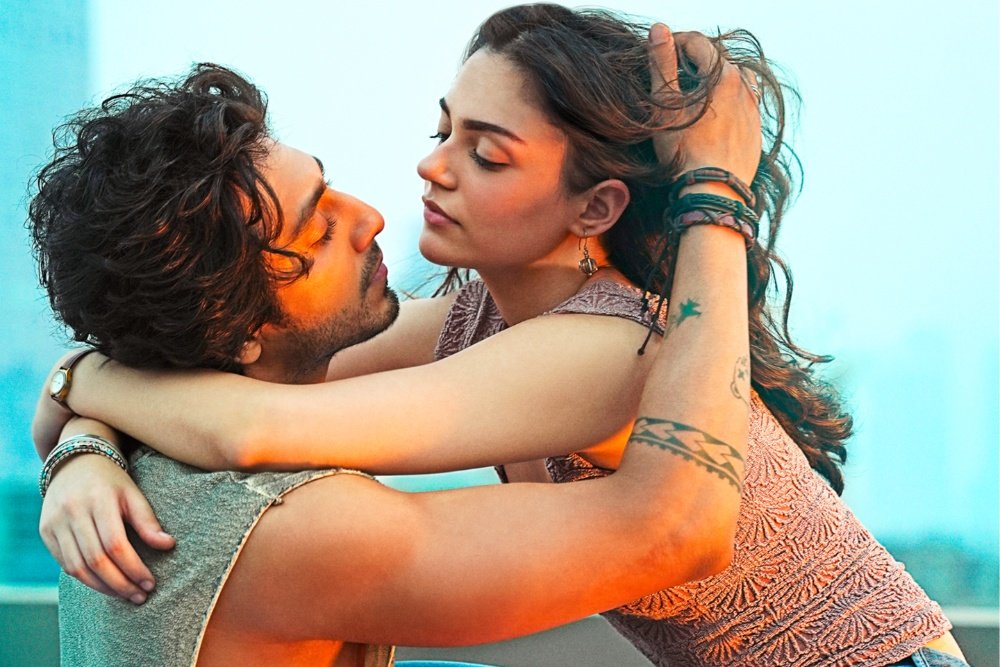 A man and woman embrace closely, foreheads nearly touching, against a blurred teal background, with the woman wearing a textured pink top and visible tattoos on her arm.