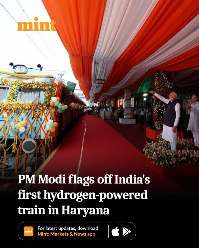 A decorated train adorned with flowers and orange, white, and green balloons sits beneath a draped canopy as a man in white attire waves beside a red carpet.