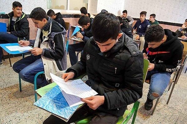 Young men in winter jackets sit at desks in a classroom, reviewing papers and writing with pens.