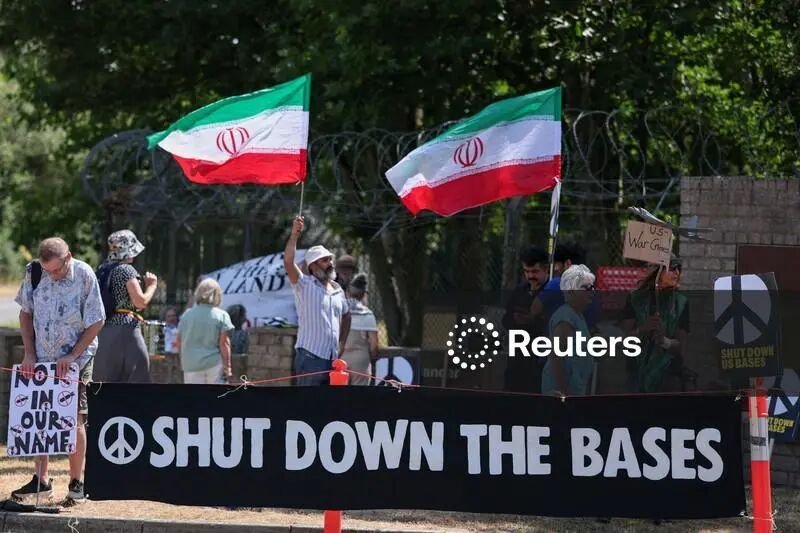 Protesters gather behind a barbed-wire fence, waving Iranian flags and holding a large banner reading "SHUT DOWN THE BASES" alongside a peace symbol.
