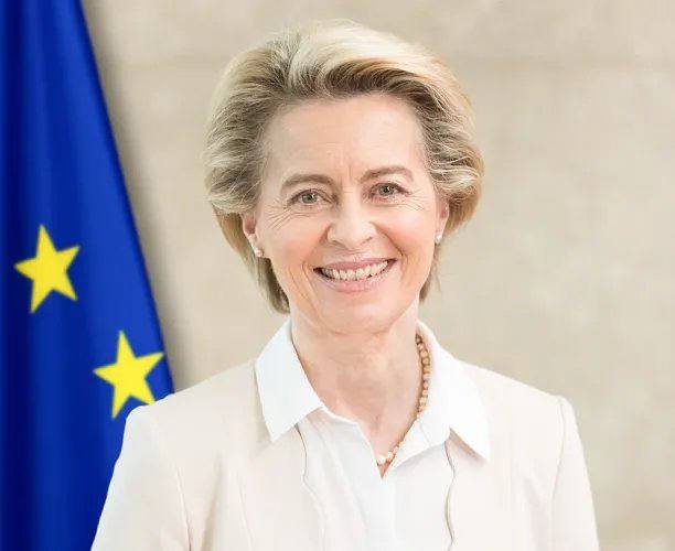 A smiling woman with short blonde hair wearing a white blouse and pearl necklace stands in front of a blue European Union flag with yellow stars.