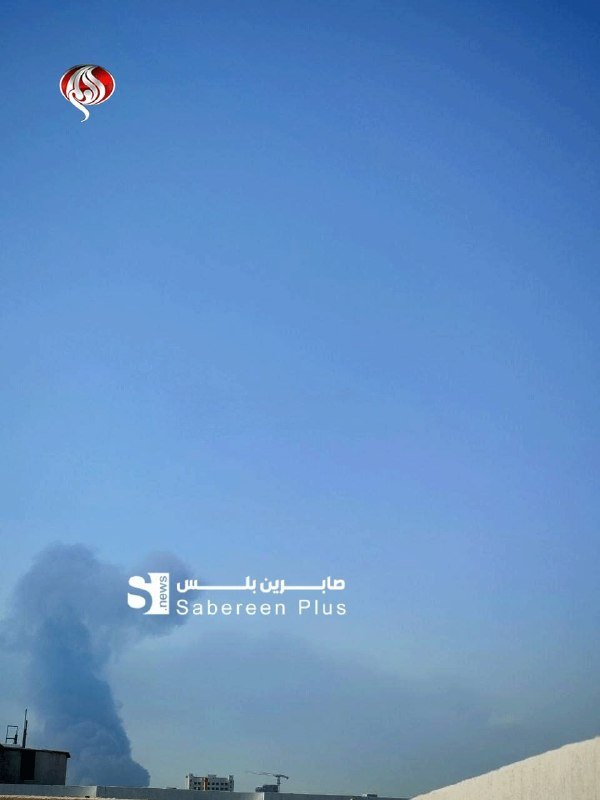 A plume of dark smoke rises from a rooftop against a clear blue sky, with a news watermark overlaid on the image.