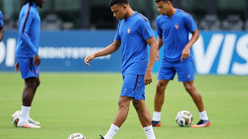 Soccer players in blue training kits walk across a grass pitch during a practice session, with soccer balls and a Visa-branded advertising board visible in the background.