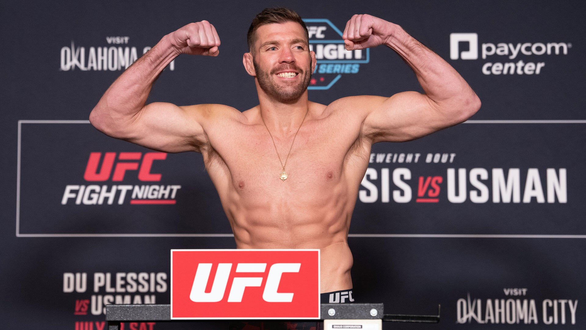 A shirtless, muscular man flexes both biceps and smiles at a UFC weigh-in event, with "UFC Fight Night" and "Paycom Center" branding visible in the background.