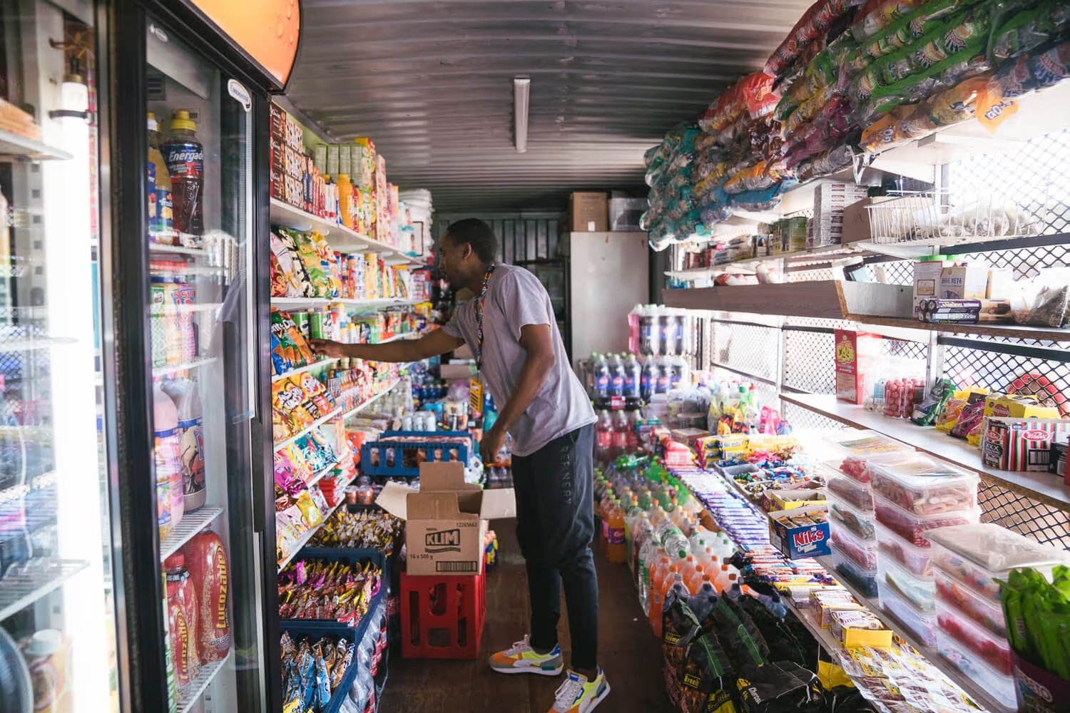A man in a gray t-shirt and dark pants reaches toward shelves of candy inside a small convenience store stocked with snacks and beverages.