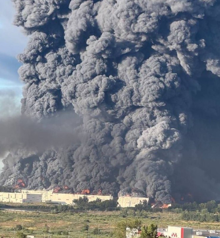 A massive plume of thick black smoke and visible flames engulf an industrial facility, rising into towering clouds against a partly cloudy sky.