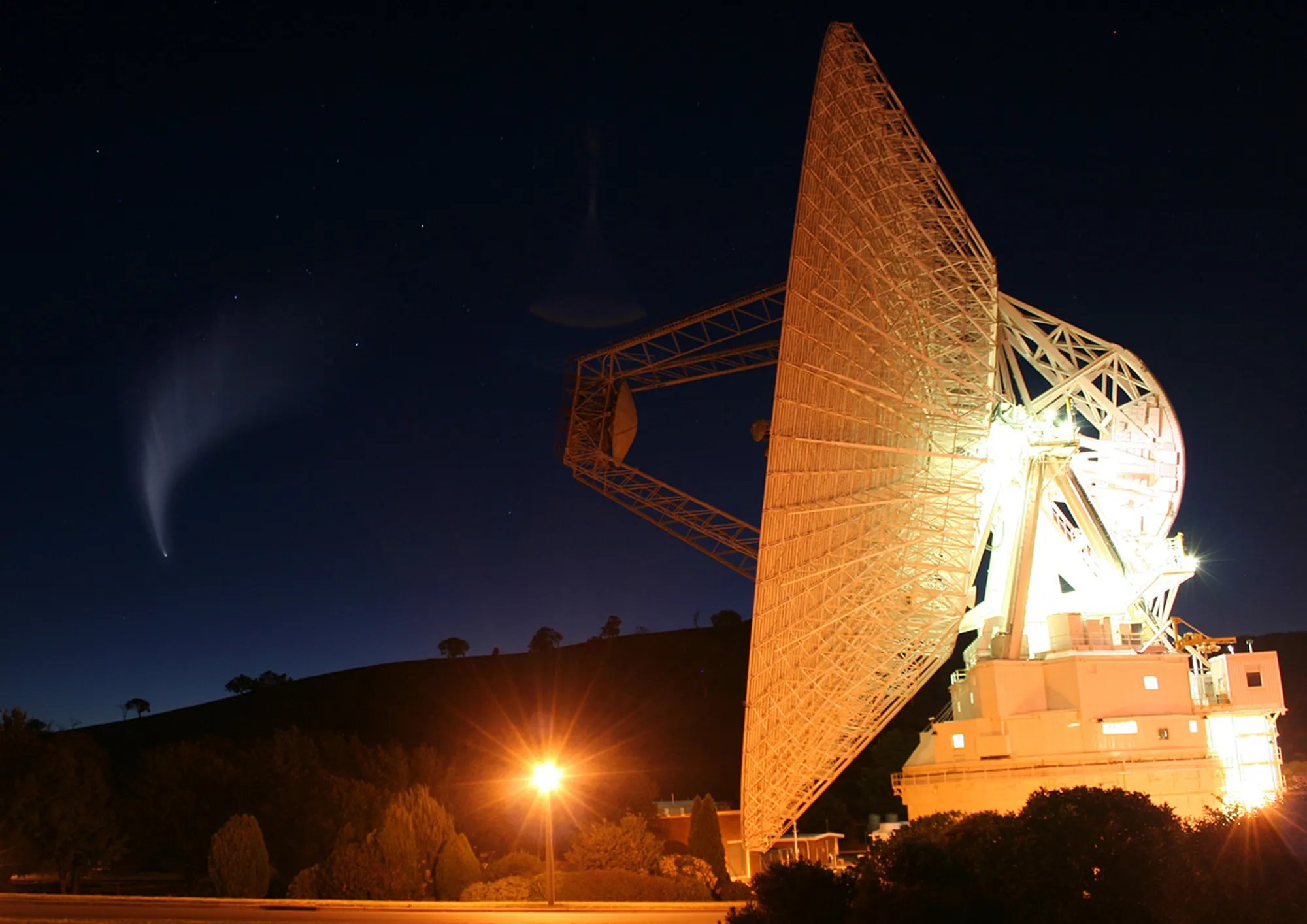 A large illuminated satellite dish antenna stands against a starry night sky, with a comet visible in the upper left and a streetlight glowing in the foreground.