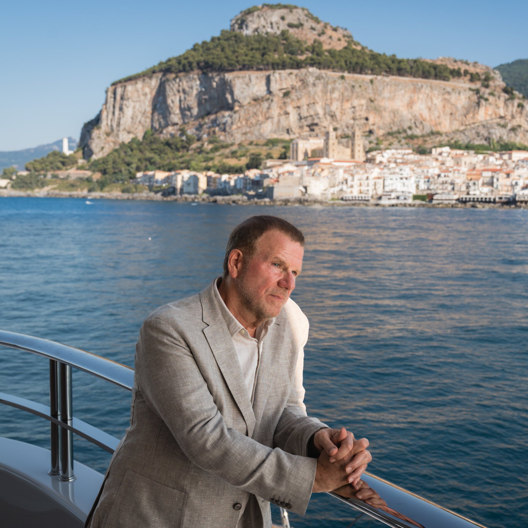 A man in a beige blazer leans against the railing of a boat, with a coastal town and large rocky mountain visible across the water behind him.