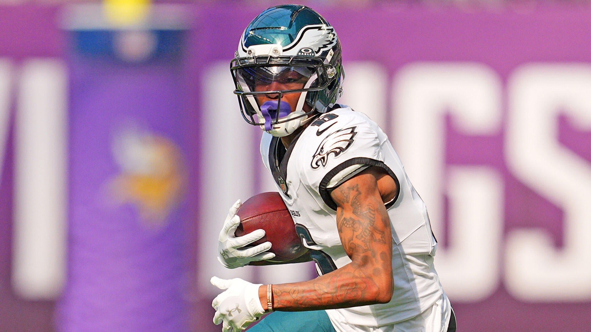 A football player in a white jersey and dark green Eagles helmet runs with the ball tucked under his arm, set against a blurred purple sideline backdrop.
