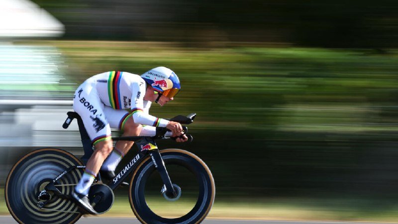 A cyclist in a rainbow-striped Bora-Hansgrohe kit rides a black time-trial bike, captured in motion against a blurred green background.