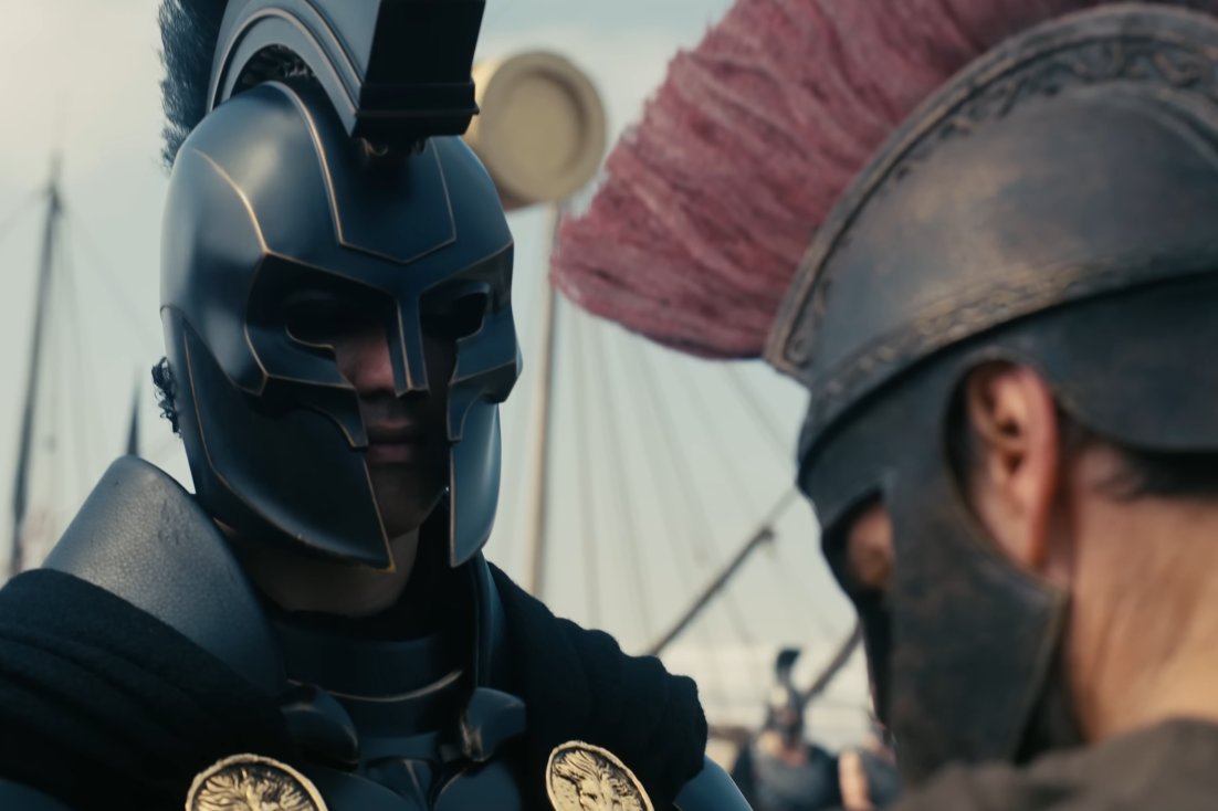 Two armored warriors face each other, one in a sleek black helmet with a crest, the other in a bronze-colored helmet with a red plume, against a backdrop of spears and shield.
