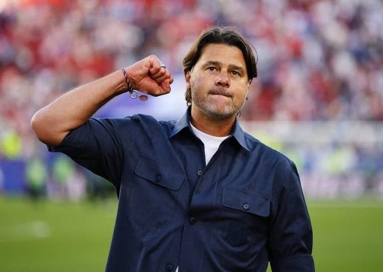 A man in a navy button-up shirt raises a clenched fist while standing on a grass field before a blurred stadium crowd.
