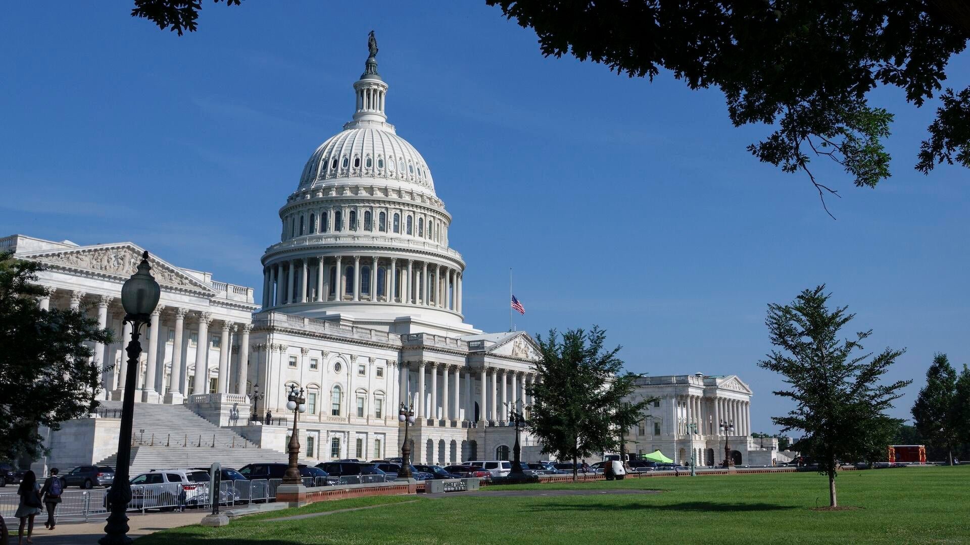 The United States Capitol building stands under a clear blue sky, its white dome and neoclassical columns visible behind parked cars, a small grassy lawn, and an American flag.