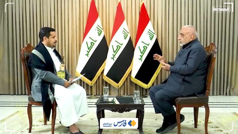 Two men sit facing each other in ornate chairs, conversing with three Iraqi flags displayed between them, while one holds a notepad and the other gestures with his hands.
