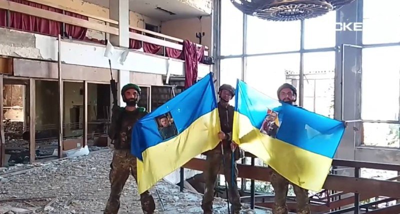 Three helmeted soldiers in camouflage uniforms stand inside a damaged building, holding large blue-and-yellow Ukrainian flags bearing printed portraits.