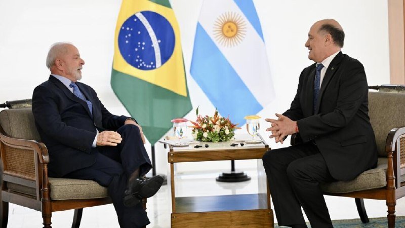 Two men in dark suits sit facing each other in armchairs, conversing with the Brazilian and Argentine flags displayed behind them and a small floral arrangement on a table between them.