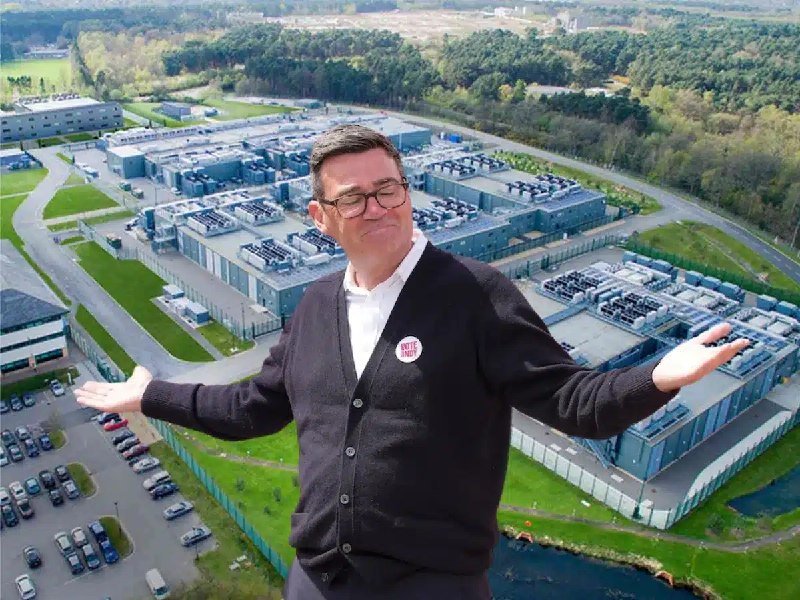 A man in glasses and a cardigan with a "Kern & Stally" button stands before an aerial view of an industrial facility with multiple buildings, parking lots, and surrounding forest.