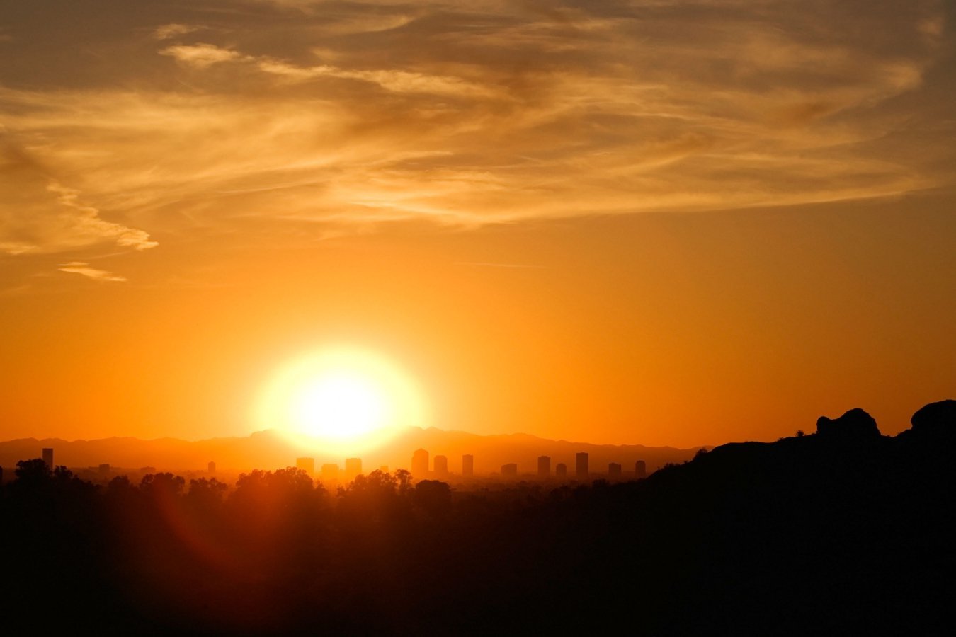 The sun sets behind a distant city skyline, casting an orange glow across a sky streaked with wispy clouds above a darkened foreground of trees and hills.