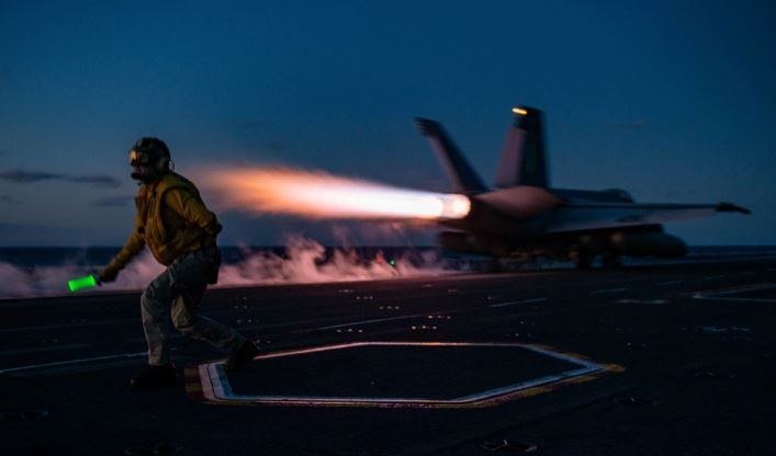 A crew member in flight gear runs across a dark flight deck while a military jet with afterburners engaged launches into a twilight sky.