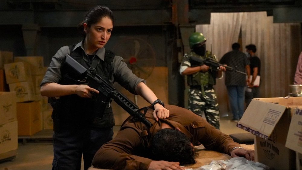 A woman holding a rifle restrains a man against a table in a warehouse, while another armed figure and onlookers stand among stacked cardboard boxes in the background.