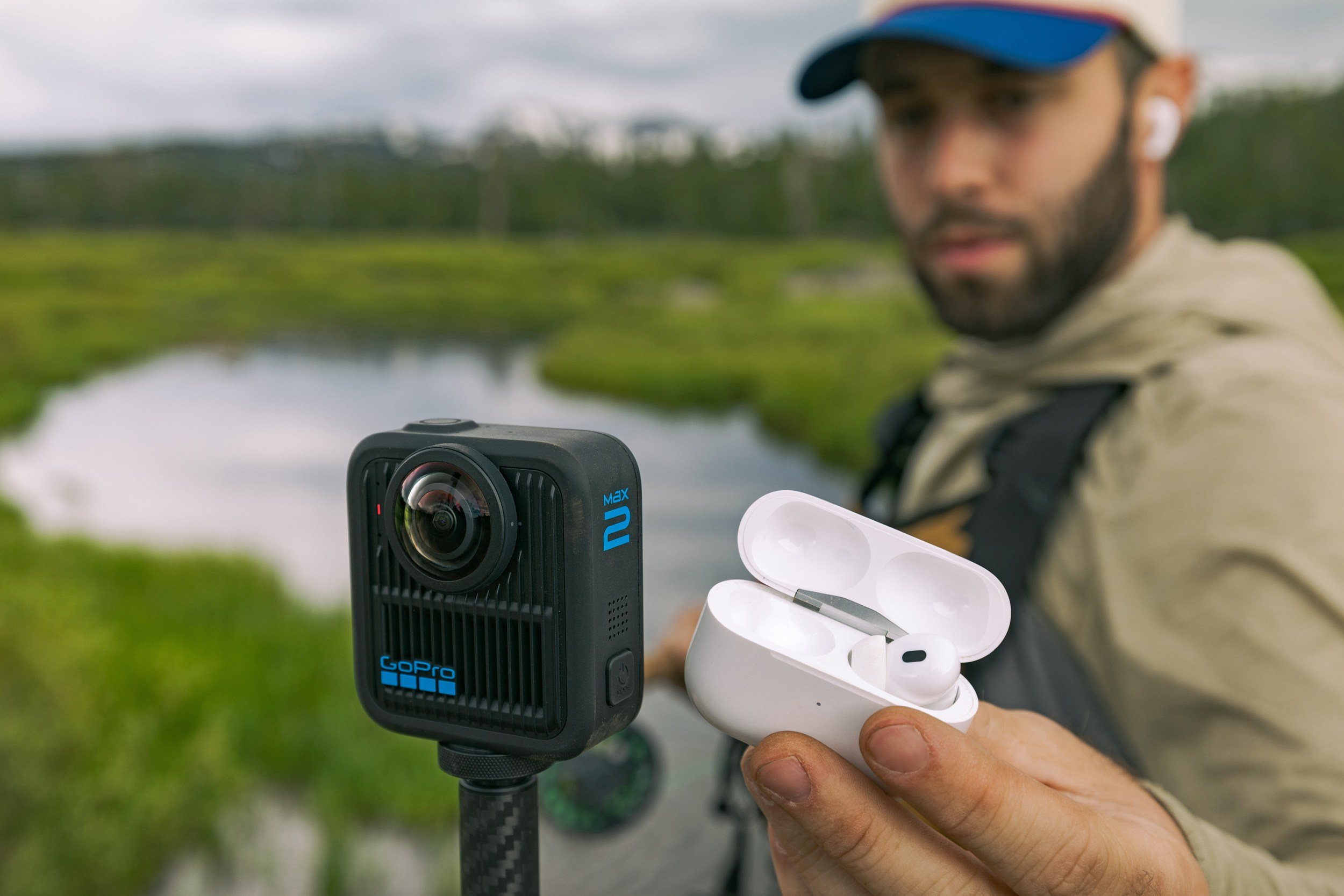 A man wearing a cap, jacket, and white earbud holds an open wireless earbud case beside a GoPro MAX camera mounted on a stick, with a marshy wetland in the background.