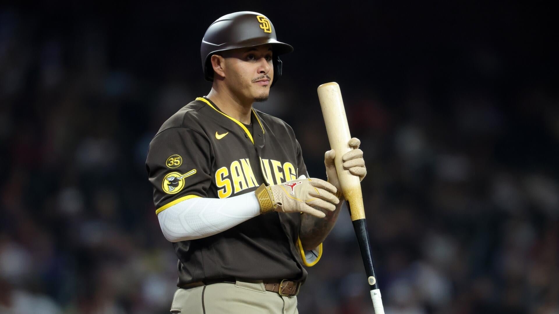 A baseball player in a brown "San Diego" jersey with the number 35 and an "SD" helmet stands holding a wooden bat, wearing batting gloves.