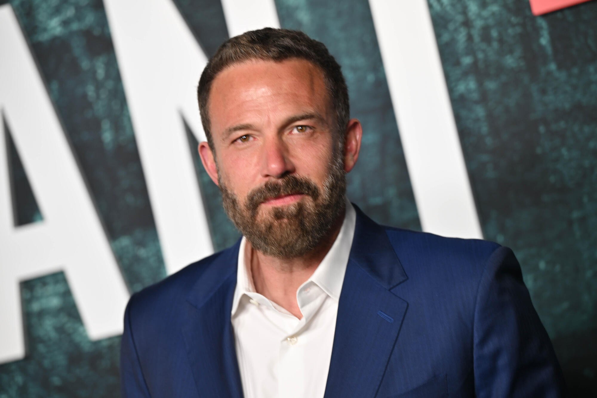A bearded man in a navy blue suit and white open-collar shirt poses in front of a dark backdrop displaying partial white and red lettering.