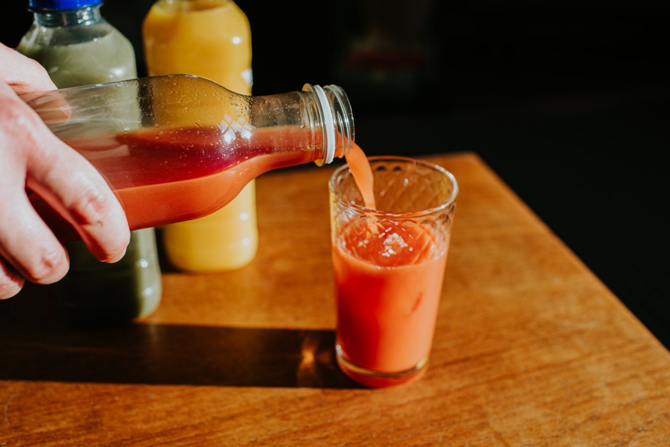 A hand pours a reddish-orange juice from a glass bottle into a clear glass on a wooden table, with two other juice bottles visible in the background.