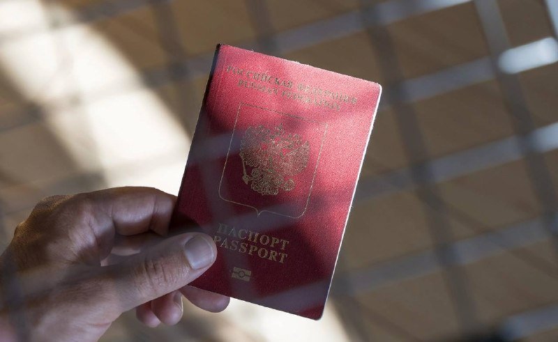 A hand holds a burgundy Russian passport featuring a gold double-headed eagle emblem, viewed through a window with a tiled floor visible in the background.